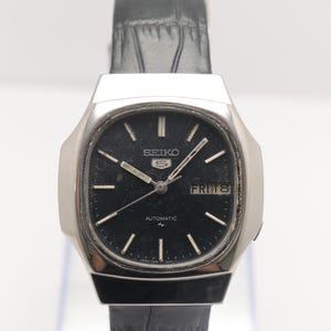 Puede incluir: Un reloj de pulsera automático Seiko vintage con esfera negra y caja plateada. El reloj muestra el día y la fecha, con una correa de cuero negro. La esfera tiene marcadores de hora y agujas plateadas.