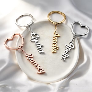 May include: Four metal keychains with custom names in cursive script. One rose gold heart-shaped ring with the name Ramsey, one silver round ring with Elyaha, one gold round ring with Amelia, and one silver heart-shaped ring with the name Joelle.