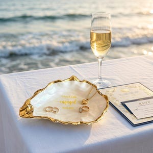 May include: A white oyster shell trinket dish with a gold-painted rim, featuring the gold text Calvin & Leila engaged 09.01.2027. Inside the dish are two gold rings and a gold heart pendant necklace. Beside it sits a champagne flute and RSVP card.