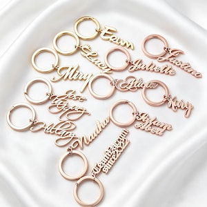 May include: A collection of metallic keyrings in gold and rose gold tones, each featuring a circular ring attached to a custom name or phrase cut out in a cursive script font. The names include Erin, Lucy, Miry, Juliette, Justino, Jose, Kary, Hillary, Nauha, Salam Abed, and Team Darym since 2022.