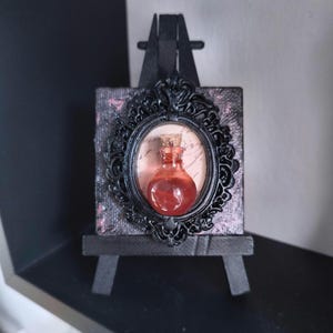 Può includere: Una piccola bottiglia di vetro riempita con un liquido rosso traslucido e sigillata con un tappo di sughero, montata all'interno di una cornice ovale nera decorata su una tela quadrata. L'oggetto è esposto su un cavalletto da tavolo nero.