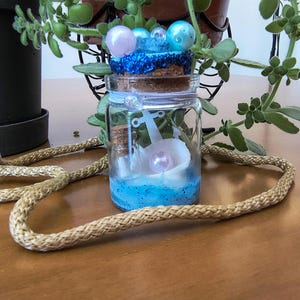 Könnte beinhalten: Ein klares Glasgefäß mit einem Korkstopfen, der mit blauem Glitzer sowie hellblauen und lavendelfarbenen Perlen verziert ist. Im Inneren befinden sich ein weißer Anker, eine weiße Muschel mit einer lavendelfarbenen Perle und blauer Glitzersand.