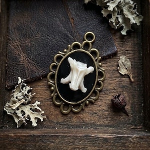 Peut inclure: Un pendentif ovale doté d'un cadre en métal couleur bronze antique avec un bord orné et festonné. Le centre contient une incrustation noire lisse, sur laquelle est monté un petit fragment d'os texturé blanc cassé rappelant une vertèbre.