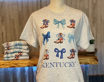 May include: A light gray t-shirt featuring a design of blue bows and cowboy boots. The word "Kentucky" is printed in blue below the design. Several folded shirts with the same design are stacked on a wooden shelf.