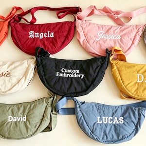 May include: A collection of nine crescent-shaped crossbody bags in various colors including orange, red, pink, gray, cream, black, yellow, olive green, and blue. Each bag features an adjustable strap and custom white or brown embroidered text.