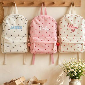 Könnte beinhalten: Drei Rucksäcke hängen an einer Holzleiste. Der linke Rucksack ist cremefarben mit einem kleinen dunklen Muster und dem Namen JAMES in Blau. Der mittlere ist rosa mit einem roten Erdbeermuster und dem Namen Daisy. Der rechte ist cremefarben mit roten Erdbeeren und dem Namen Naomi.