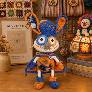 May include: A handmade crochet bunny doll with blue, orange, and white accents. The doll has large ears, button details, and a cape. It is decorated with charms and bells. The background includes framed art and other crochet items.