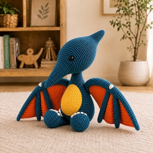 May include: A handmade blue crochet pterodactyl plush toy with orange and yellow accents. The wings are orange with white details. The toy has a long beak and a yellow belly. The plush is sitting on a neutral-colored rug.