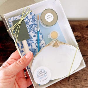 Op de afbeelding: Een doorzichtige doos bevat een mini-schelpen decoupagekit. De kit bevat een witte schelp, Mod Podge, een penseel, een schuimborstel, een blauw-wit bloemenpatroon servet en een klein verfpotje. De kit is vastgebonden met een gouden lint.