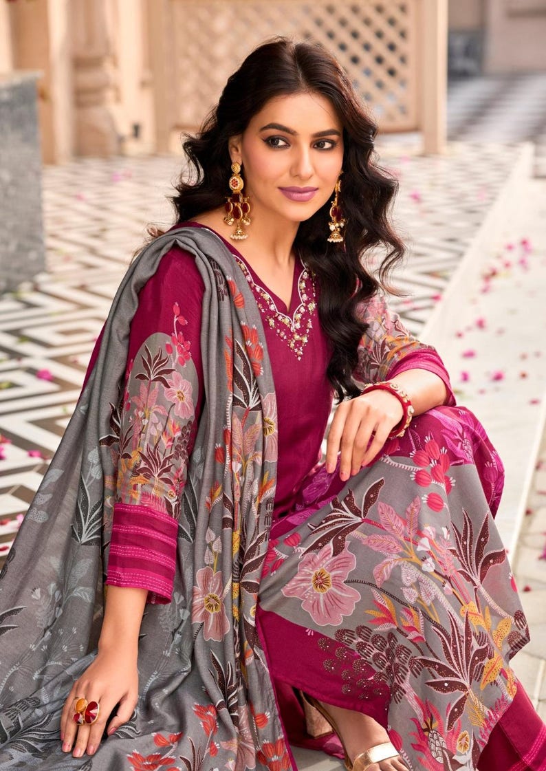 May include: A magenta long-sleeved tunic with a V-neckline featuring white embroidery, paired with a matching magenta pant and a grey shawl. The shawl is decorated with a large-scale floral print in shades of pink, orange, and dark green.
