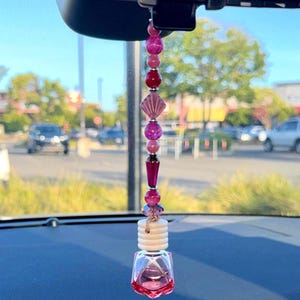 Könnte beinhalten: Ein Auto-Lufterfrischer, der am Rückspiegel hängt, mit einer vertikalen Reihe aus rosa, magentafarbenen und dunkelroten Perlen in verschiedenen Formen und Texturen, gekrönt von einer weißen Holzkappe und einem facettierten, transparenten rosa Glasflakon.