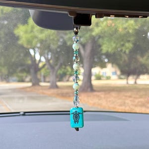 May include: A car rearview mirror hanging ornament featuring a vertical strand of iridescent, light green, and teal beads. The bottom of the strand features a rectangular turquoise-colored bead with a black turtle graphic printed on the front.