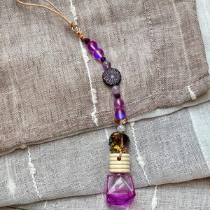Può includere: Un diffusore di oli essenziali viola con una catena di perline decorativa. La catena presenta perline viola, iridescenti e trasparenti, una perlina floreale nera e viola e un anello di legno. Il diffusore è una piccola bottiglia di vetro sfaccettata.