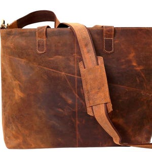 May include: A brown leather tote bag with a shoulder strap and top handles. The bag has a rectangular shape and a distressed finish. The shoulder strap is adjustable and padded for comfort. The bag is designed for carrying items.