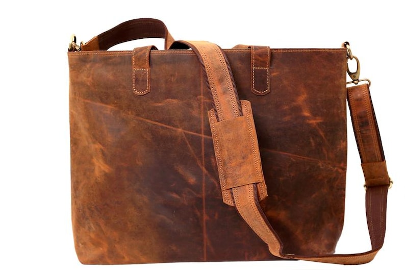 May include: A brown leather tote bag with a shoulder strap and top handles. The bag has a rectangular shape and a distressed finish. The shoulder strap is adjustable and padded for comfort. The bag is designed for carrying items.