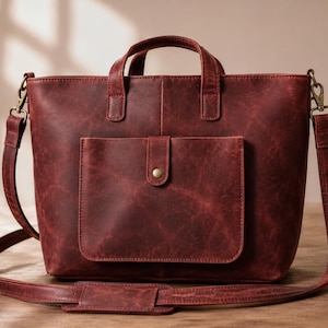 May include: A burgundy leather tote bag with a front pocket and a shoulder strap. The bag has two top handles and a flap pocket with a brass button closure. The leather has a distressed look.