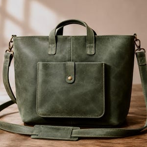 May include: Olive green leather tote bag with a front pocket and a top handle. The bag has a detachable shoulder strap and a gold-colored button closure on the front pocket. The bag is sitting on a wooden surface.