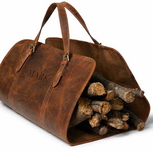 Könnte beinhalten: Ein brauner Leder-Holzträger mit zwei Griffen und dem eingeprägten Namen "MARK" auf der Seite. Der Träger ist mit Brennholz gefüllt. Das Leder hat einen Used-Look.