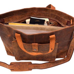 May include: Open brown leather tote bag with a front pocket and shoulder strap. The bag is filled with items, including a passport and files. The bag has a distressed look.