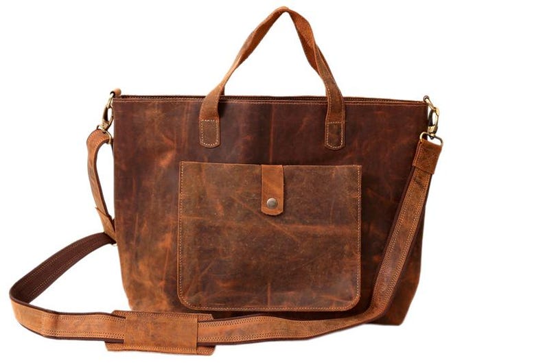 May include: A brown leather tote bag with a front pocket and a shoulder strap. The bag has two top handles and a detachable shoulder strap. The leather has a distressed look, with variations in color and texture. The bag is designed for carrying items.