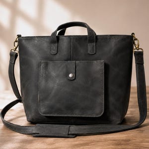 May include: A black leather tote bag with a top handle and a detachable shoulder strap. The bag features a front pocket with a button closure and gold-colored hardware. The bag is sitting on a wooden surface.