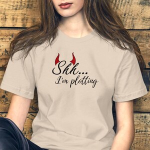 Puede incluir: Camiseta beige con la frase "Shh... I'm plotting" en negro. Encima del texto, cuernos de diablo rojos. La camiseta de manga corta tiene cuello redondo.