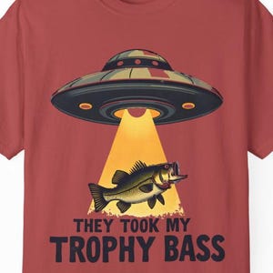 May include: A brick red t-shirt featuring a graphic of a grey UFO hovering above a green bass fish. The UFO emits a yellow beam of light onto the fish. Below the graphic, the text reads "THEY TOOK MY TROPHY BASS" in bold, dark blue capital letters.