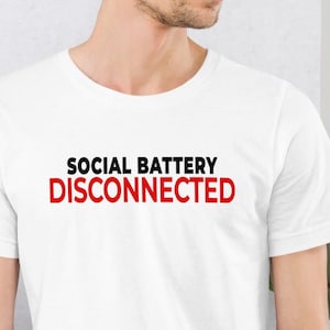 Peut inclure: T-shirt blanc avec l'inscription "SOCIAL BATTERY DISCONNECTED" en noir et rouge. Le texte est centré sur le devant du t-shirt. Le col est rond et le tissu semble doux.