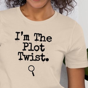 Puede incluir: Camiseta beige claro con la frase "I'm The Plot Twist." en negro con fuente goteante. Debajo, un pequeño icono de lupa. Una prenda casual para el uso diario.