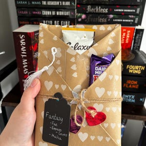 May include: A brown paper envelope with a white heart pattern containing a Galaxy chocolate bar and a Cadbury Dairy Milk bar. A black tag labelled Fantasy, Fae, Enemies, Lovers, Mafia is attached with twine, along with two small red and purple hearts.