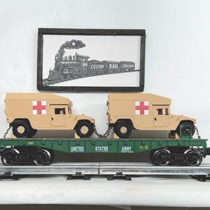 May include: A green United States Army flatbed train car carrying two tan military ambulance vehicles with red cross symbols. Above the train car is a framed black and white sign featuring a steam locomotive illustration and the text Custom Rail Creations.