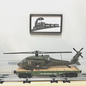 May include: An olive green military helicopter model secured to a flatbed train car with the text UNITED STATES ARMY printed on the side. Above the train car, a framed black and white sign depicts a steam locomotive with the text CUSTOM RAIL CREATIONS.