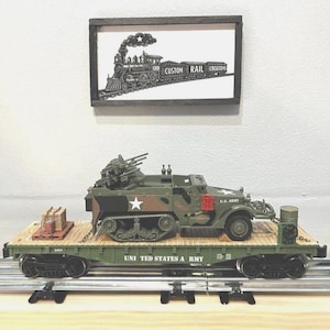 May include: A model flatcar train carriage in olive green with the text UNITED STATES ARMY, carrying a camouflaged military half-track vehicle with a mounted gun turret and a small wooden crate. Above, a framed sign depicts a steam locomotive.