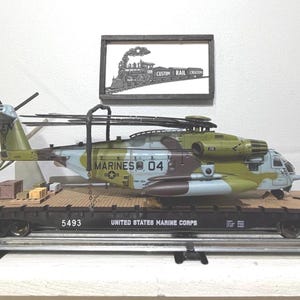 May include: A model helicopter with a camouflage paint scheme labeled MARINES 04 is secured to a flatbed train car marked UNITED STATES MARINE CORPS 5493. Above the train car, a framed sign displays a black and white steam locomotive illustration with the text CUSTOM RAIL CREATIONS.