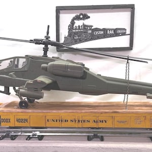 May include: A model olive green military attack helicopter secured by chains onto a yellow flatbed train car. The train car features the text UNITED STATES ARMY and the number 40224. A framed sign above displays a black and white train illustration.