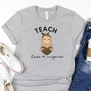 May include: A heather gray t-shirt with the words "TEACH love inspire" and a cartoon sloth design. The sloth has a leopard print heart and headband. White sneakers and blue jeans are also in the image.