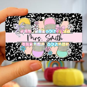 May include: A rectangular card featuring a black and white composition notebook paper design. The card has colorful pencil illustrations with bows and flowers. A pink banner across the center reads "Mrs. Smith" in black script.