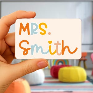 May include: A rectangular name tag with rounded corners, held in a hand. The tag is cream-colored with the words "MRS. Smith" in orange, yellow, and blue script. The background is a classroom setting with colorful furniture and a rainbow.