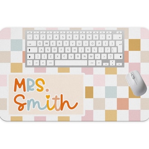May include: A rectangular desk mat with a colorful checkered pattern in shades of pink, yellow, and white. A white keyboard and mouse are on the mat. The mat has the text "MRS. Smith" in colorful lettering.