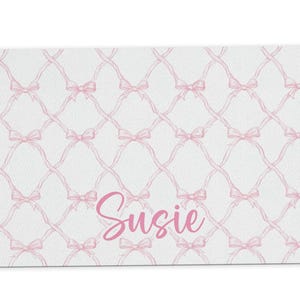 May include: A white doormat with a pink bow pattern and the name "Susie" written in pink.