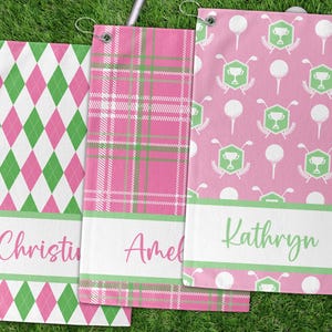 May include: Three golf towels with different patterns and names. One has a pink, white, and green argyle design, another has a pink and green plaid pattern, and the third has a pink background with golf-themed icons. Each towel has a white band with a name.