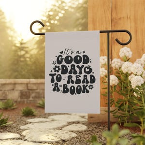 May include: A white garden flag with black text that reads "It's a good day to read a book." The text is in a retro font with flowers and stars surrounding it.