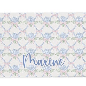 May include: A white bath mat with a blue and pink floral pattern and the name "Maxine" written in blue.