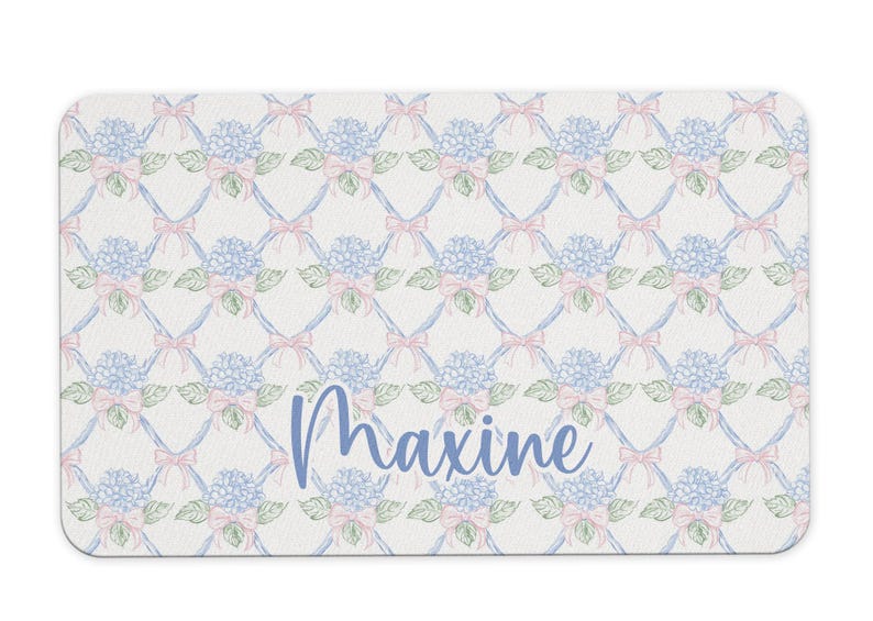 May include: A white bath mat with a blue and pink floral pattern and the name "Maxine" written in blue.