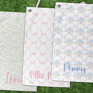 May include: Three personalized golf towels with different patterns and names. One towel has a floral pattern with the name "Lexi" in pink. Another features a pink bow design with the name "Ellie M" in pink. The third towel has a floral pattern with the name "Penny" in blue.