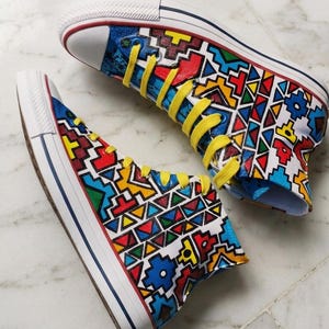 May include: High-top canvas sneakers with a vibrant, geometric pattern in red, yellow, blue, and green. The shoes have white soles, toe caps, and laces, with yellow laces. The design covers the entire shoe, creating a bold and colorful statement.