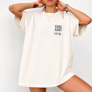 Puede incluir: Una camiseta oversize de color blanco roto con el texto negro COOL AUNT club impreso en la parte superior izquierda del pecho. La prenda tiene mangas cortas, cuello redondo y un corte holgado con un diseño gráfico sencillo.