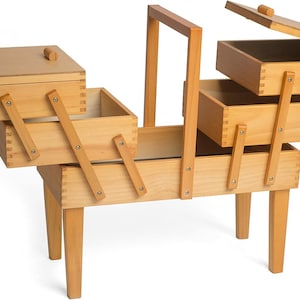 May include: A light-coloured wooden sewing box featuring a cantilevered design with multiple tiered storage compartments. The box stands on four legs, includes a central carrying handle, and has hinged lids that open to reveal internal storage trays.