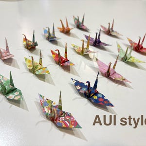 May include: A collection of origami paper cranes in various colors and patterns, including pink, green, blue, and yellow. The cranes are arranged on a white surface. Text in the image reads "AUI style".