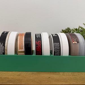 May include: A green rectangular organizer holding six cylindrical containers with black and white labels. The containers are filled with various products. The word "SPART" is visible on one of the labels. The organizer sits on a wooden surface.